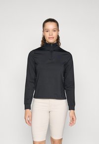 Crna atletska zip-up majica s dugim rukavima i visokim ovratnikom. Ima glatku teksturu i uski kroj. Nosiva uz svijetlosmeđe šortse.