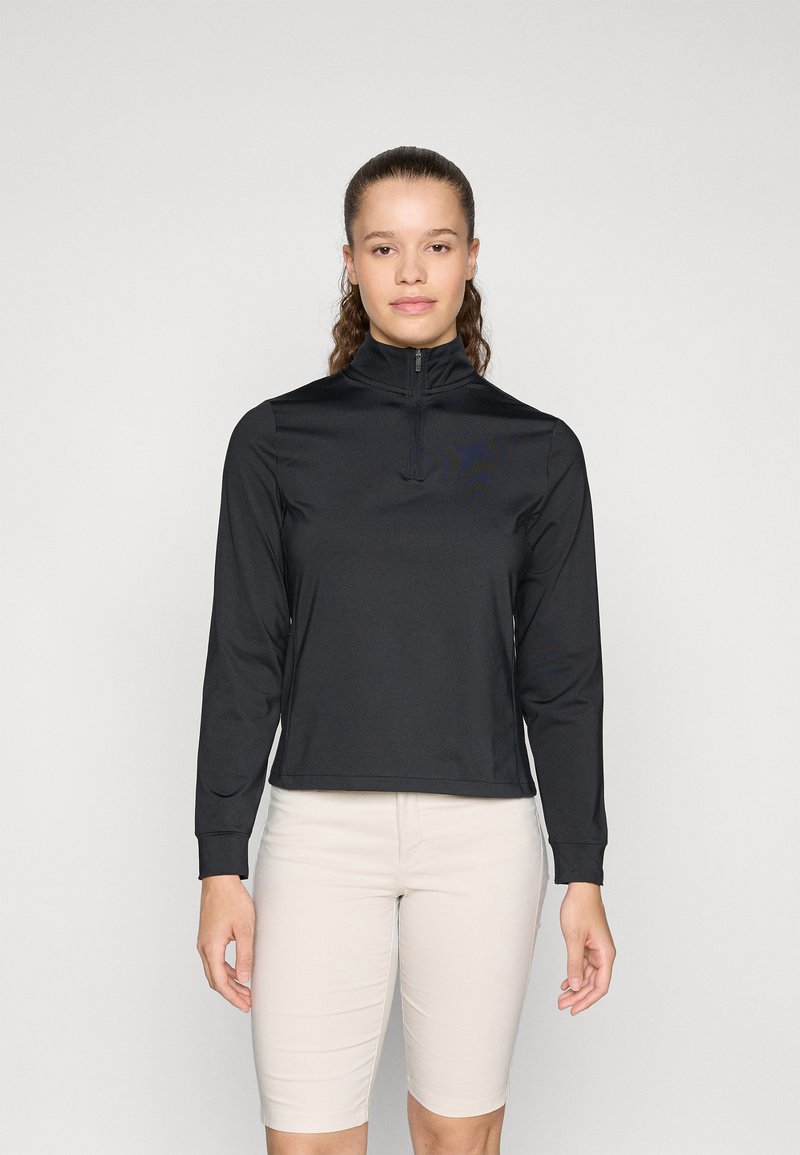 Crna atletska zip-up majica s dugim rukavima i visokim ovratnikom. Ima glatku teksturu i uski kroj. Nosiva uz svijetlosmeđe šortse.