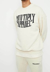 Sudadera color crema hecha de una tela suave, con un atrevido texto negro "MULTIPLY APPAREL" en el frente. Diseño holgado con ribetes acanalados.