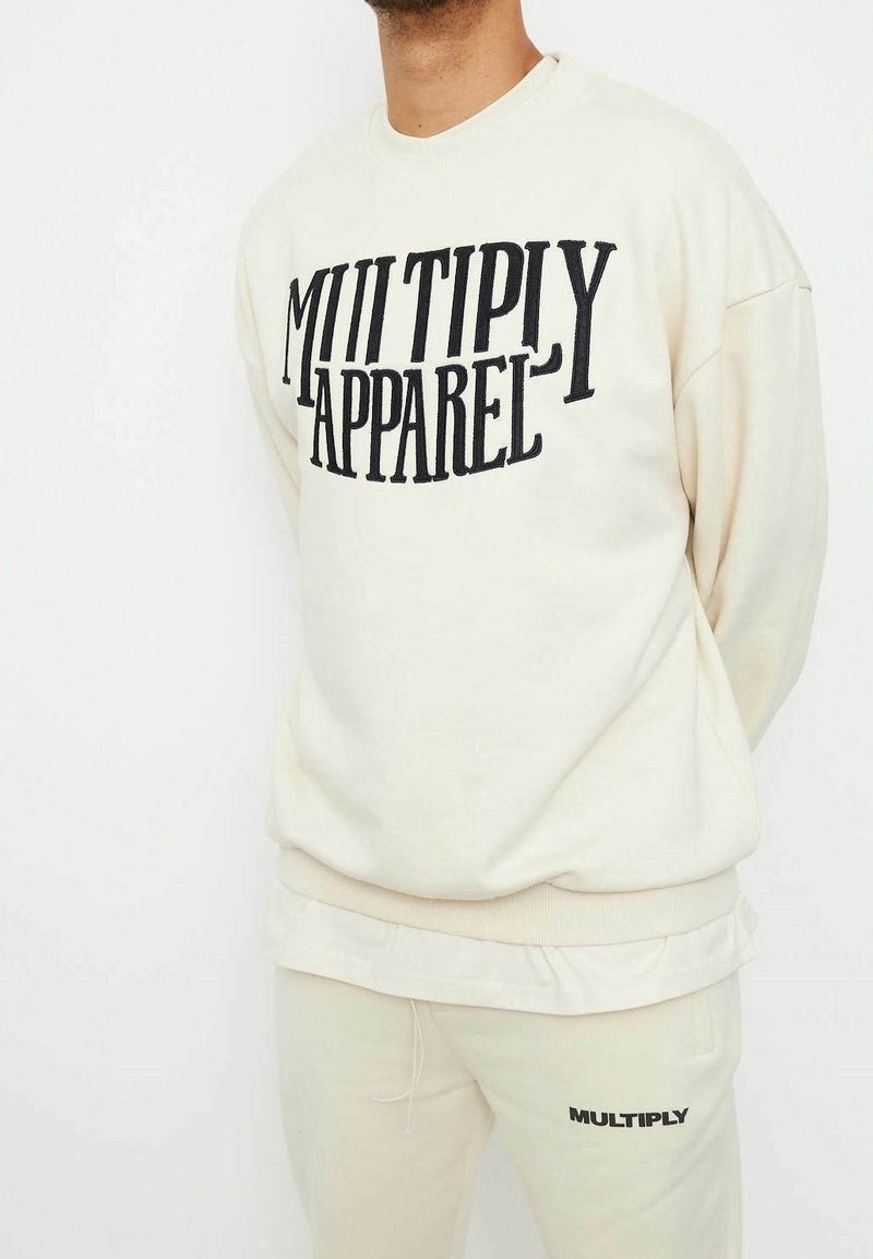Sudadera color crema hecha de una tela suave, con un atrevido texto negro "MULTIPLY APPAREL" en el frente. Diseño holgado con ribetes acanalados.