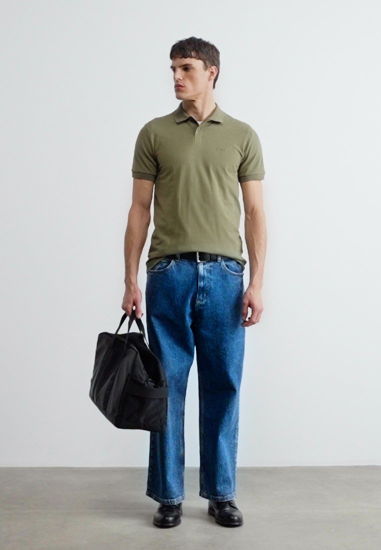 Jeune homme debout tenant un sac de sport noir, portant un polo vert olive, un jean bleu et des chaussures noires, sur fond blanc uni.