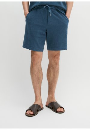 Mężczyzna noszący niebieskie szorty z sznurkiem i ciemnobrązowe skórzane klapki, stojący z rękami w kieszeniach na jednolitym tle.