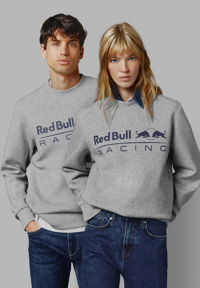 Sudadera gris con el logo de "Red Bull Racing" en azul marino, con puños y dobladillo acanalados, combinada con jeans de mezclilla azul oscuro.