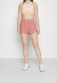 Sujetador deportivo en melocotón con patrón geométrico; pantalones cortos rosas con cintura elástica y logo de Nike en el lateral. Zapatillas deportivas blancas.