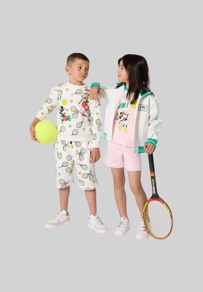 Due bambini in abiti a tema tennis che tengono una grande palla da tennis e una racchetta da tennis, in piedi davanti a uno sfondo semplice.