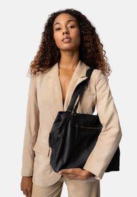 Sort læderhåndtaske med en blød tekstur, der har en lynlås-lomme og guldfarvede hardware-detaljer. Båret af en person i et lysebeige outfit.