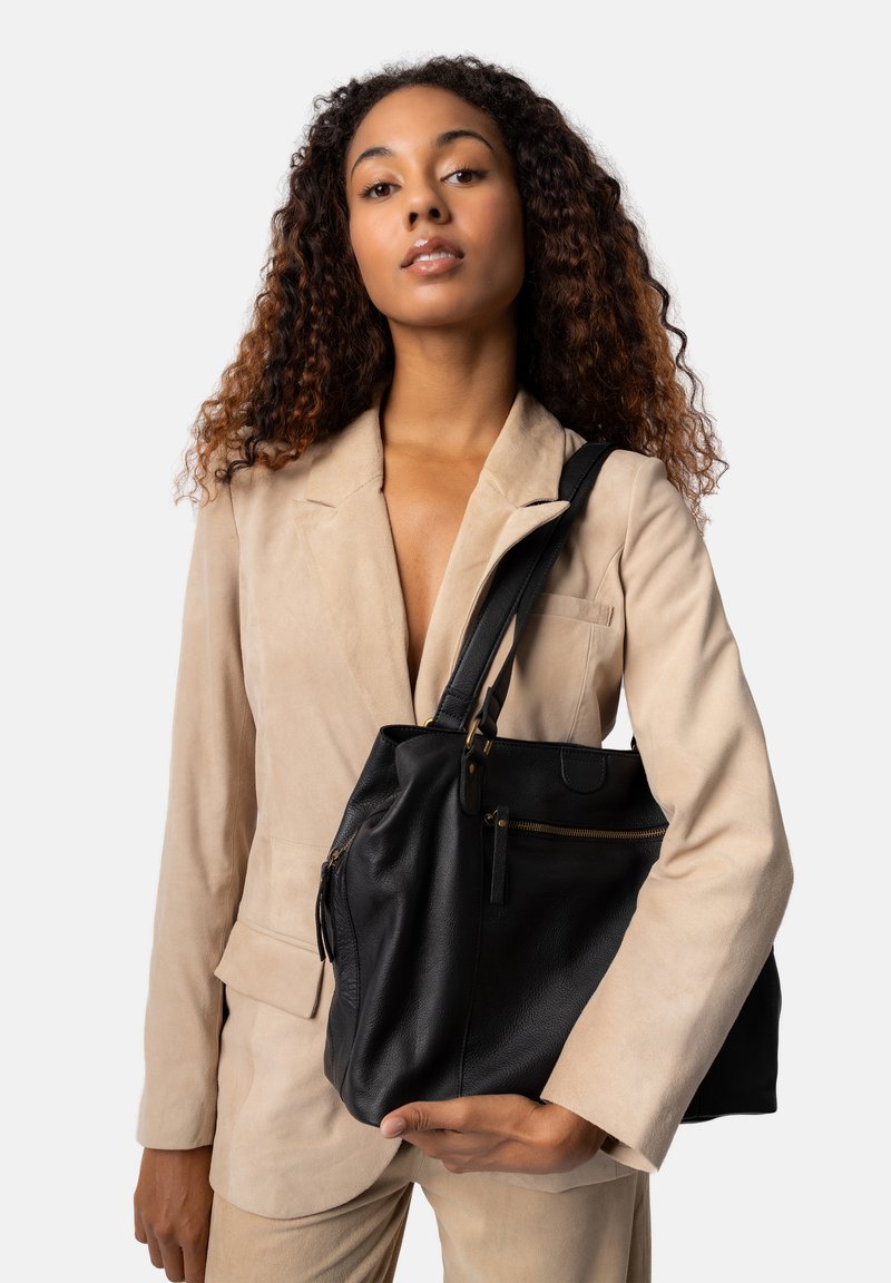 Sort læderhåndtaske med en blød tekstur, der har en lynlås-lomme og guldfarvede hardware-detaljer. Båret af en person i et lysebeige outfit.