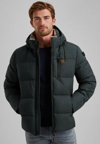 Veste rembourrée vert foncé avec une fermeture éclair à l'avant, une capuche et une doublure douce. Comprend une poche sur la poitrine et des détails en tissu texturé.