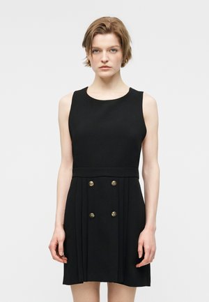 Kobieta o krótkich jasnobrązowych włosach, ubrana w czarną sukienkę bez rękawów z czterema złotymi guzikami na plisowanej spódnicy, stoi na tle białego tła.