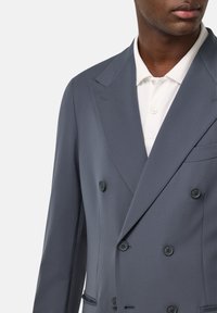 Sivina dvoredna blazer jakna z ostrimi reverji in dvema gumboma. Prirejen dizajn, gladka tekstura in en sam žep na prsih. Nosi se čez belo polo majico.
