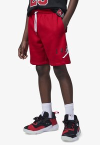 Pantalones cortos de algodón rojos con cordón blanco. Presenta un gráfico de "JORDAN" y una silueta de baloncesto. Combinado con zapatillas deportivas negras y rojas y calcetines blancos.