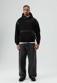 Zwarte hoodie met een voorzak, voorzien van een roze logo. Draagt losse, donkergrijze jeans en zwarte schoenen met roze veters.