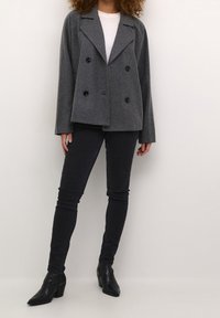 Grau meliertes, zweireihiges Wollmischjackett mit breitem Revers, zwei Fronttaschen und schwarzen Knöpfen, kombiniert mit dunklen Jeans und schwarzen Stiefeletten.