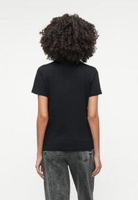 Melna kokvilnas T-krekls ar īsām piedurknēm, piegulīgas formas, gluda tekstūra un klasiskā apaļā kakla izgriezums. Valkāts ar izbalējušām pelēkām džinsām.