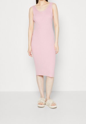 Woman wearing a sleeveless, ribbed, light pink midi dress with beige frayed slide sandals standing against a plain white background.