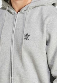 Gray zip-up hoodie made of cotton blend, featuring a white drawstring, ribbed cuffs, and small black Adidas logo on the left chest.