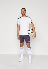 Jersey blanco de manga corta con rayas negras en los hombros, pantalones cortos morados, calcetines blancos con rayas azules y botas blancas, sosteniendo un balón de fútbol.