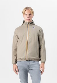 Chaqueta cortavientos beige claro con capucha, que presenta una cremallera frontal con franjas amarillas, naranjas y azules, y puños elásticos.