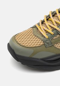 Scarpa da ginnastica verde e beige con tomaia in rete, suola in gomma e lacci gialli. I punti di texture sul materiale aggiungono interesse visivo.