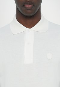 Hvit polo-shirt med strukturert veving, med en tre-knappslukking og en liten heksagonal logo på venstre bryst.