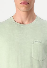Világos zöld pamut póló, kerek dekoltázzsal, rövid ujjakkal és mellzsebbel, amelyen hímezett "Barbour" részlet található. Puha tapintású.