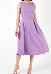 Robe midi sans manches lavande avec corsage ajusté et jupe plissée, portée avec des sandales à talons hauts et bride à la cheville assorties en lavande.