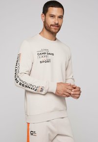 Lys grå sweatshirt lavet af blødt stof, med grafisk tekst i sort og orange, samt ribbede manchetter og talje.