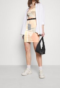 Kolorowa sukienka bodycon w pastelowych odcieniach z geometrycznymi wzorami, w połączeniu z oversize'ową białą koszulą i małą czarną torebką. Białe sneakersy.