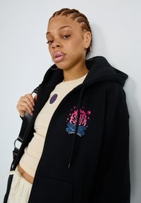 Svart hettegenser med et frontgrafikk av et leirbål i rosa og blått, med snor og kengurulomme. Hatt over en beige topp.