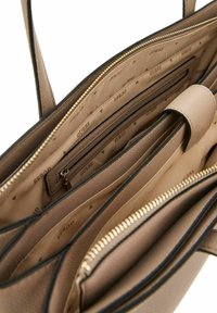 Sac à main tan avec une surface texturée, des compartiments zippés et une doublure beige présentant un motif de logo subtil. Des sangles ajustables sont incluses.
