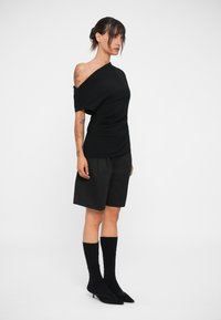 Top nero asimmetrico con drappeggi, abbinato a pantaloni short neri su misura. Il modello indossa calze nere fino al ginocchio e décolleté neri a punta.