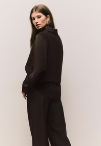 Brown cropped sweater over a sheer shirt, paired with wide-leg pants. Smooth texture with a subtle sheen; features a collar and buttoned cuffs.