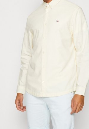 Man draagt een crèmekleurig overhemd met knopen en een klein Tommy Hilfiger-logo op de borst, gecombineerd met lichtblauwe broek.