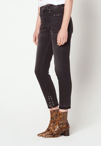 Personne portant un jean skinny noir avec des détails œillet à la cheville et des bottines marron à motif peau de serpent, debout devant un fond uni.