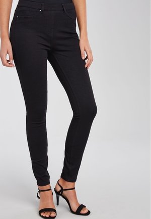 Kobieta ubrana w dopasowane czarne spodnie oraz czarne sandały na wysokim obcasie, stojąca na tle jednolitego, jasnoszarego tła.