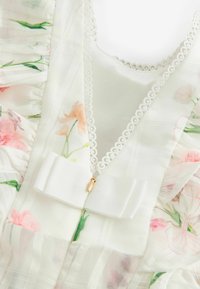 Transparente, floral gemusterte Stoff mit cremigem Grund, der mit Rüschen, einem mit Spitze eingefassten Ausschnitt und einer Schleifen-Applikation am Reißverschluss geschmückt ist.