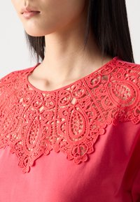 Haut en corail avec un décolleté en dentelle complexe aux motifs de goutte d'eau ; texture de tissu lisse ; design à col arrondi ; silhouette ajustée.