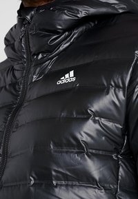 Schwarze gepolsterte Jacke mit Reißverschluss, Kapuze und dem Adidas-Logo in Weiß auf der Vorderseite. Glattes, glänzendes Material mit genähten Abschnitten.