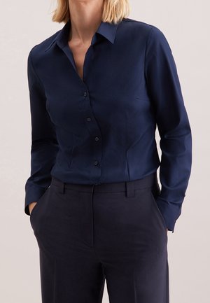 Persoon die een donkerblauw overhemd met knopen draagt, ingestopt in bijpassende donkerblauwe broek, met handen in de zakken, tegen een lichte achtergrond.