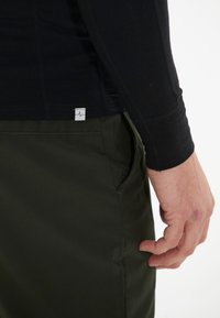 Zwarte langemouwen top met een aansluitend ontwerp, gecombineerd met donkergroene broek met een zijzak en een klein wit label met een hartslagpictogram.