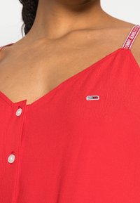 Top sin mangas rojo con escote en V, que presenta un frente abotonado y un pequeño logo bordado en el lado izquierdo del busto.