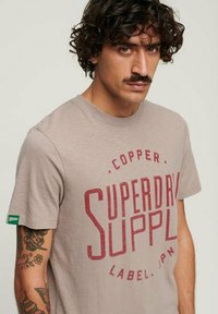 Camiseta de algodón gris con un atrevido estampado gráfico en rojo "SUPERDRY SUPPLY", mangas cortas y un pequeño logo verde en la manga izquierda.