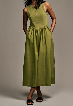 Woman wearing sleeveless olive green midi dress with gathered waist and slip-on black sandals, standing with hands in pockets.