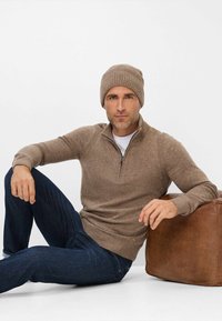 Brauner gestrickter Pullover mit einem Viertelreißverschluss, über einem weißen T-Shirt getragen, kombiniert mit dunklen Jeans und einer passenden braunen Mütze.