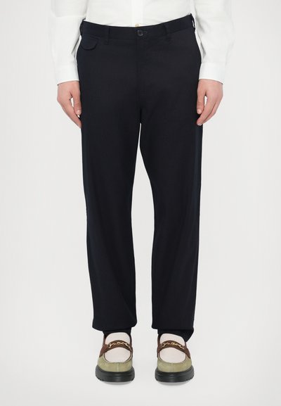 Pantaloni neri con una vestibilità sartoriale, tessuto liscio, tasche laterali e passanti per cintura, abbinati a scarpe multicolori con un accento hardware prominente.