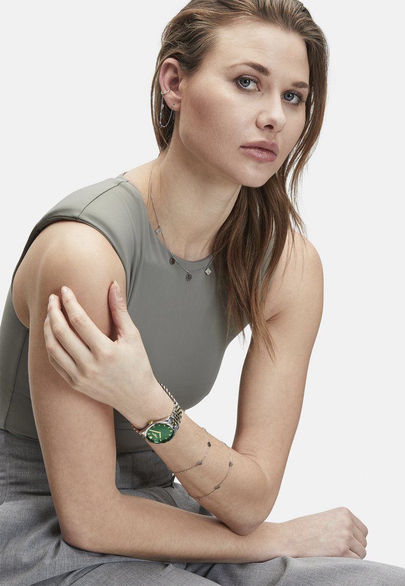 Montre verte avec un cadran rond et un bracelet argenté, superposée avec des bracelets en chaîne argentés. Le modèle porte un haut gris ajusté, mettant en valeur les bijoux.