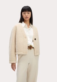 Chaqueta de forro polar beige con corte holgado, botones en el frente y dos bolsillos. Llevada sobre una camisa blanca y pantalones beiges, mostrando un estilo casual en capas.