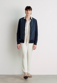 Chaqueta bomber acolchada en color navy con puños de canalé, combinada con una camisa crema y pantalones a juego, completada con sandalias marrones. Diseño simple y moderno.