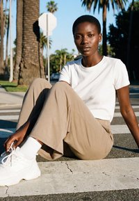 Rövid hajú nő fehér pólóban, bézs nadrágban és fehér tornacipőben, aki egy utcai járdaszegélyen ül, háttérben pálmafákkal.