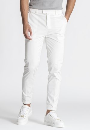 Muž v úzkych bielych nohaviciach, bielych teniskách a svetlosivom rebrovanom tričku stojaci s rukami čiastočne vreckách.